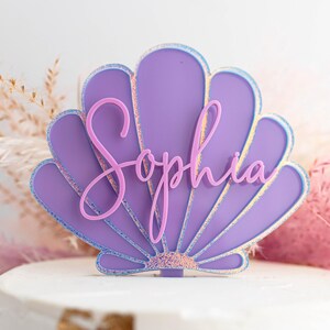 Pu&ograve; includere: Un topper per torta a forma di conchiglia viola con il nome Sophia in un carattere corsivo rosa. La conchiglia presenta un bordo iridescente scintillante e una base testurizzata, con un piccolo stelo per l'inserimento nella torta.