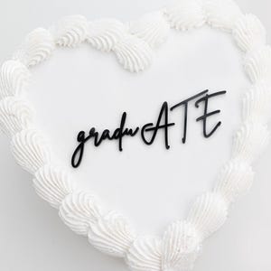 May include: A heart-shaped cake with white frosting and a border of piped white frosting. The word "graduate" is written in black script across the center. A celebratory dessert for a graduation party.