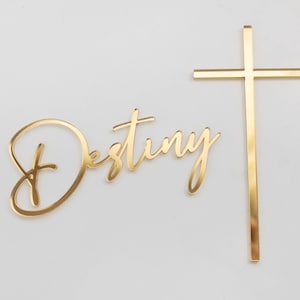 May include: A gold-colored wall decoration featuring the word Destiny in a cursive script font positioned to the left of a simple, vertical Christian cross. Both elements have a smooth, reflective, metallic finish and are presented together.