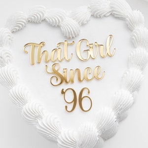 May include: A white heart-shaped cake with gold lettering that reads "That Girl Since '96".