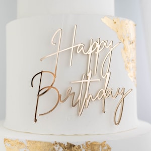 May include: A gold-colored acrylic cake topper featuring the words Happy Birthday in a cursive script font. The topper is placed on a white frosted cake decorated with gold leaf accents on the side, creating a reflective and metallic appearance.