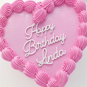 May include: A heart-shaped cake with smooth pink frosting and a glittery finish. The edges are decorated with piped pink frosting swirls. Centered on the top is white cursive text that reads Happy Birthday Linda in a clean, script-style font.
