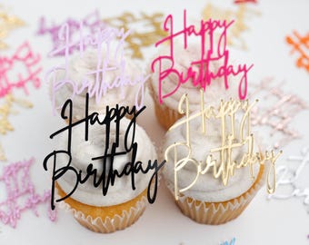 May include: Four cupcakes with white frosting, each topped with a Happy Birthday cake topper in different colors including light pink, hot pink, black, and gold. Additional Happy Birthday cutouts in various colors are scattered around the cupcakes.