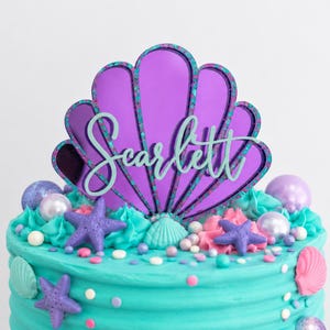 Pu&ograve; includere: Un topper per torta a forma di conchiglia a specchio viola con bordo verde acqua e il nome Scarlett in corsivo verde acqua. Il topper &egrave; posizionato su una torta con glassa color turchese decorata con stelle marine viola, conchiglie e perle.
