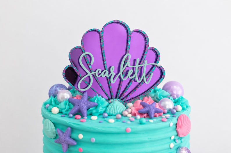Pu&ograve; includere: Un topper per torta a forma di conchiglia a specchio viola con bordo verde acqua e il nome Scarlett in corsivo verde acqua. Il topper &egrave; posizionato su una torta con glassa color turchese decorata con stelle marine viola, conchiglie e perle.