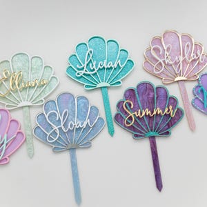 Pu&ograve; includere: Una collezione di sette decorazioni per torte a forma di conchiglia in vari colori, tra cui menta, verde acqua, rosa, blu, viola e viola scuro. Ogni decorazione presenta un nome unico in corsivo, come Elliana, Lucian, Skylar, Lily, Sloan, Summer e Natalia.