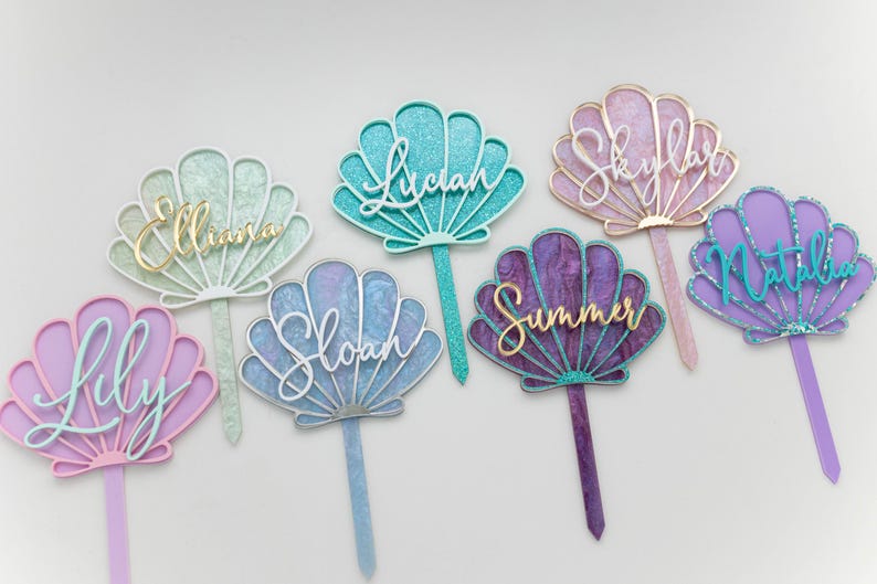 Pu&ograve; includere: Una collezione di sette decorazioni per torte a forma di conchiglia in vari colori, tra cui menta, verde acqua, rosa, blu, viola e viola scuro. Ogni decorazione presenta un nome unico in corsivo, come Elliana, Lucian, Skylar, Lily, Sloan, Summer e Natalia.