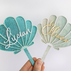 Pu&ograve; includere: Due topper per torta a forma di conchiglia con bastoncini. Il topper a sinistra &egrave; color ottanio glitterato con il nome Lucian in corsivo bianco. Il topper a destra &egrave; verde pallido marmorizzato con il nome Elliana in corsivo oro.