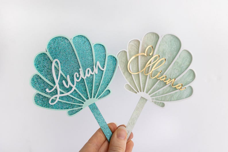 Pu&ograve; includere: Due topper per torta a forma di conchiglia con bastoncini. Il topper a sinistra &egrave; color ottanio glitterato con il nome Lucian in corsivo bianco. Il topper a destra &egrave; verde pallido marmorizzato con il nome Elliana in corsivo oro.
