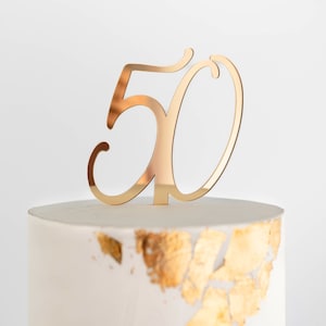 May include: Gold mirror-like cake topper in the shape of the number 50. The number is in a cursive font.