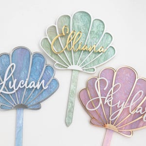 Pu&ograve; includere: Tre topper per torta a forma di conchiglia con superfici in acrilico marmorizzato blu, verde e rosa. Ogni topper presenta un nome in corsivo: Lucian in bianco, Elliana in oro e Skylar in bianco, con un gambo abbinato per l'inserimento.