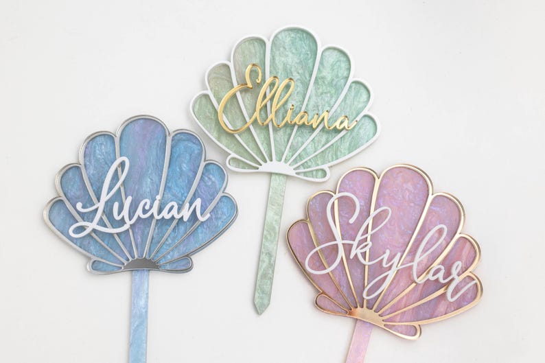 Pu&ograve; includere: Tre topper per torta a forma di conchiglia con superfici in acrilico marmorizzato blu, verde e rosa. Ogni topper presenta un nome in corsivo: Lucian in bianco, Elliana in oro e Skylar in bianco, con un gambo abbinato per l'inserimento.