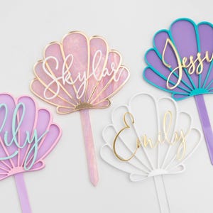 Pu&ograve; includere: Quattro decorazioni per torte a forma di conchiglia con picchetti appuntiti. Una &egrave; rosa con il testo Skylar in bianco, una viola e turchese con il testo Jessie in oro, una lilla con il testo Lily in azzurro e una bianca con il testo Emily in oro.