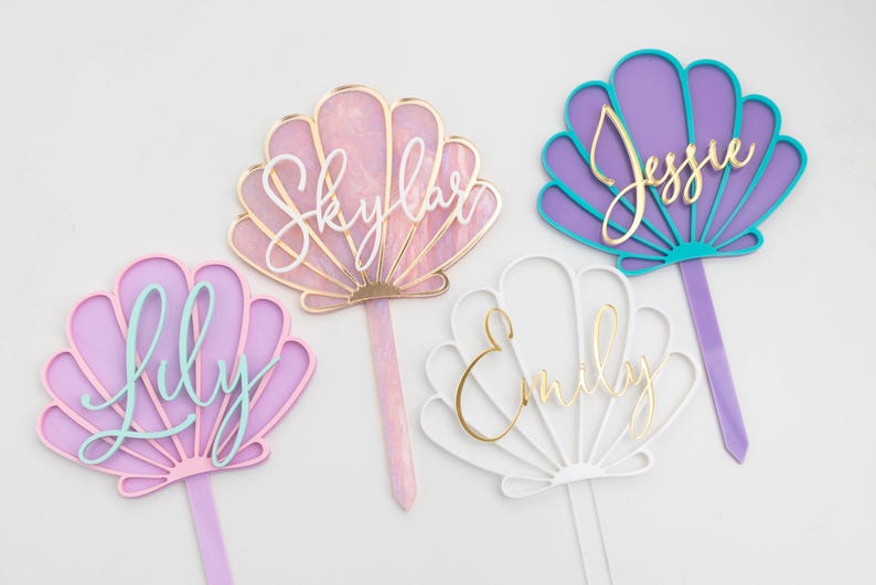 Pu&ograve; includere: Quattro decorazioni per torte a forma di conchiglia con picchetti appuntiti. Una &egrave; rosa con il testo Skylar in bianco, una viola e turchese con il testo Jessie in oro, una lilla con il testo Lily in azzurro e una bianca con il testo Emily in oro.