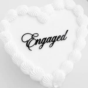 May include: A heart-shaped cake with white frosting and a decorative border of piped frosting. The word "Engaged" is written in black script across the top of the cake. Perfect for an engagement celebration.