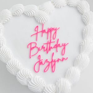 May include: A heart-shaped cake with smooth white frosting and a decorative piped border. Centered on the top is a bright pink script cake topper that reads Happy Birthday Jazlynn in a cursive font, standing out against the white surface.