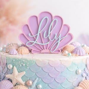 Pu&ograve; includere: Un topper per torta a forma di conchiglia color lavanda con il nome Lily scritto in corsivo azzurro. Il topper &egrave; posizionato su una torta rotonda decorata con squame da sirena color pastello, stelle marine bianche e perle decorative.