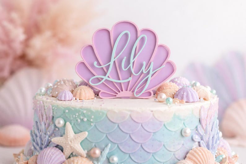 Pu&ograve; includere: Un topper per torta a forma di conchiglia color lavanda con il nome Lily scritto in corsivo azzurro. Il topper &egrave; posizionato su una torta rotonda decorata con squame da sirena color pastello, stelle marine bianche e perle decorative.