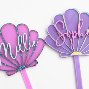 Pu&ograve; includere: Due decorazioni per torta a forma di conchiglia in acrilico. Quella a sinistra &egrave; rosa con un bordo glitterato color ottanio e il nome Millie in corsivo azzurro. Quella a destra &egrave; viola con un bordo dorato e il nome Sophia in corsivo rosa.