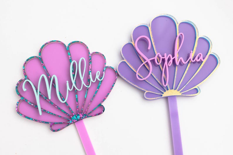 Pu&ograve; includere: Due decorazioni per torta a forma di conchiglia in acrilico. Quella a sinistra &egrave; rosa con un bordo glitterato color ottanio e il nome Millie in corsivo azzurro. Quella a destra &egrave; viola con un bordo dorato e il nome Sophia in corsivo rosa.