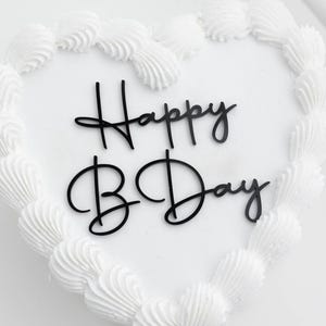 May include: A heart-shaped cake with smooth white frosting and a decorative piped border. A black script cake topper is placed in the center of the cake, featuring the text Happy B Day in a stylized, cursive font against the white surface.