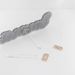 May include: A silver glitter heart-shaped sign with a clear acrylic stand. The sign is made of cardboard and has a pink glitter accent. The stand is made of two pieces of clear acrylic that are connected by a small piece of gold metal.