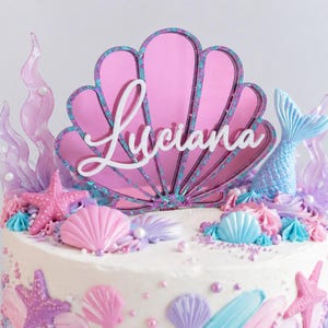 Pu&ograve; includere: Un topper per torta con una grande conchiglia in acrilico rosa con il nome Luciana in corsivo bianco. Il topper &egrave; circondato da forme di alghe viola traslucide e una coda di sirena azzurra, il tutto posizionato su una torta glassata bianca.