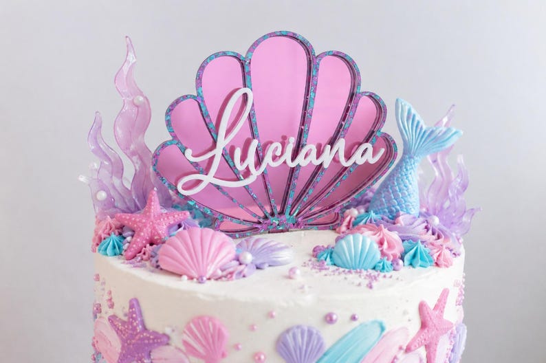 Pu&ograve; includere: Un topper per torta con una grande conchiglia in acrilico rosa con il nome Luciana in corsivo bianco. Il topper &egrave; circondato da forme di alghe viola traslucide e una coda di sirena azzurra, il tutto posizionato su una torta glassata bianca.