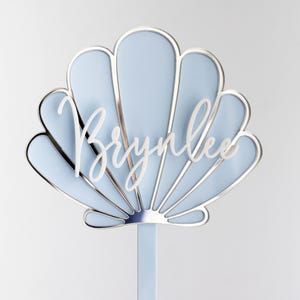 Pu&ograve; includere: Un topper per torta a forma di conchiglia azzurro con un bordo metallico argentato e il nome Brynlee scritto in corsivo bianco al centro. Il topper &egrave; fissato a un singolo bastoncino verticale azzurro nella parte inferiore centrale.