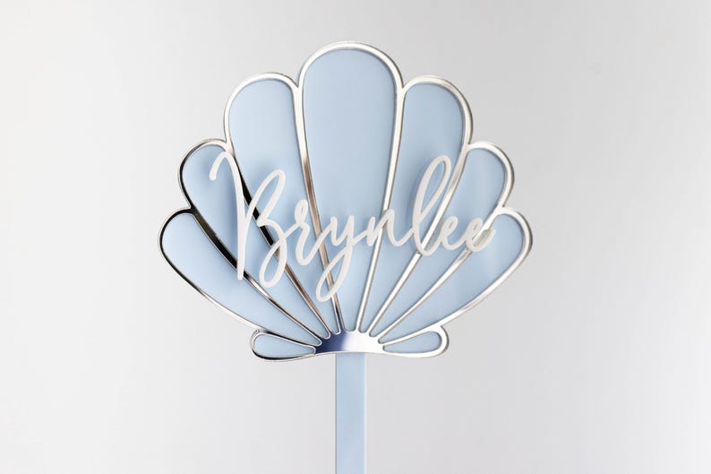 Pu&ograve; includere: Un topper per torta a forma di conchiglia azzurro con un bordo metallico argentato e il nome Brynlee scritto in corsivo bianco al centro. Il topper &egrave; fissato a un singolo bastoncino verticale azzurro nella parte inferiore centrale.