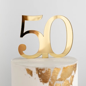 May include: A reflective gold-colored cake topper shaped as the number 50, featuring a smooth, metallic finish. The topper is positioned upright on top of a white cake decorated with gold leaf accents along the bottom edge of the visible portion.