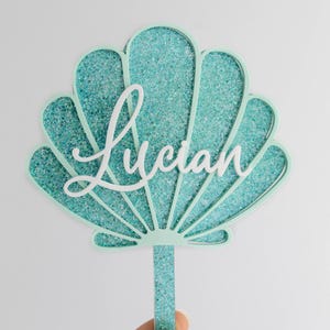 Pu&ograve; includere: Un topper per torta a forma di conchiglia con una cornice verde menta chiaro e un inserto glitterato color turchese. Il nome Lucian &egrave; scritto al centro in corsivo bianco e il topper include un gambo abbinato ricoperto di glitter.