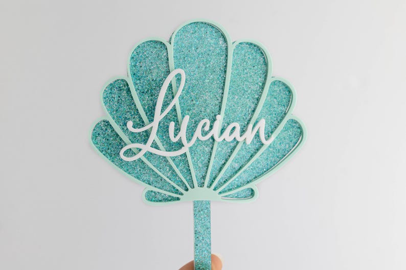 Pu&ograve; includere: Un topper per torta a forma di conchiglia con una cornice verde menta chiaro e un inserto glitterato color turchese. Il nome Lucian &egrave; scritto al centro in corsivo bianco e il topper include un gambo abbinato ricoperto di glitter.