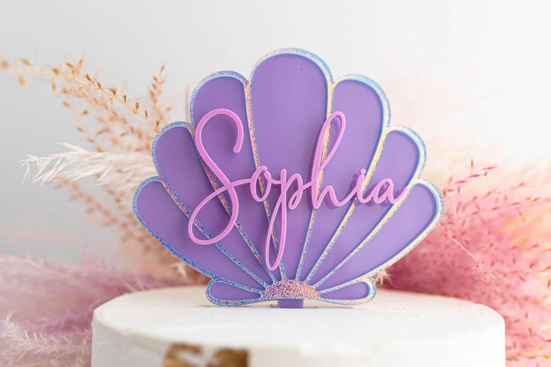 Pu&ograve; includere: Un topper per torta a forma di conchiglia viola con il nome Sophia in un carattere corsivo rosa. La conchiglia presenta un bordo iridescente scintillante e una base testurizzata, con un piccolo stelo per l'inserimento nella torta.