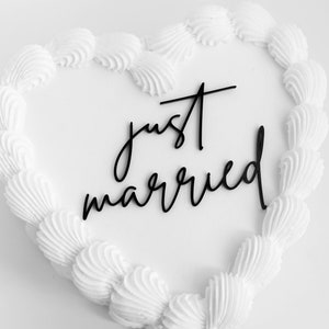 May include: White heart-shaped cake with white frosting and the words "just married" written in black script.
