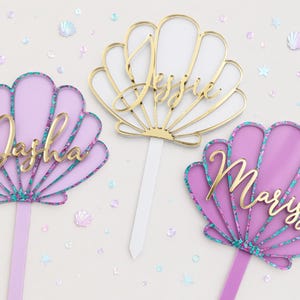 Pu&ograve; includere: Tre topper per torta a forma di conchiglia con stecchi appuntiti. Due sono color lavanda con bordi glitterati color ottanio e i nomi Dasha e Marissa in corsivo dorato. Il topper centrale &egrave; dorato con il nome Jessie e uno stecco bianco.