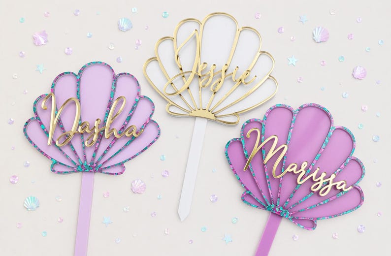 Pu&ograve; includere: Tre topper per torta a forma di conchiglia con stecchi appuntiti. Due sono color lavanda con bordi glitterati color ottanio e i nomi Dasha e Marissa in corsivo dorato. Il topper centrale &egrave; dorato con il nome Jessie e uno stecco bianco.