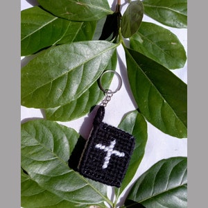 May include: A black crocheted keychain shaped like a book with a white cross on the front. The keychain is attached to a silver key ring.