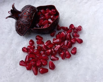 May include: A red, ornate, pomegranate-shaped container with a lid, filled with red pomegranate seeds. The container is open and some seeds have spilled out onto a white surface.