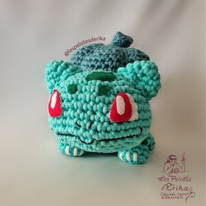 May include: A crocheted Bulbasaur plush toy in green, white, and red. The toy is sitting on a white surface. The Instagram handle @lespelotesderika is visible in the background.