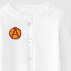 May include: A white cardigan sweater with a round neckline and long sleeves. The sweater has a button-up front and a circular patch with a yellow border and a red letter "A" on the left chest. The sweater is made of a soft knit material.