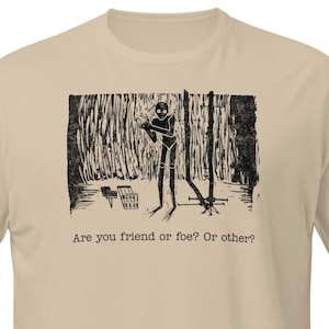 Puede incluir: Camiseta beige de manga corta con un estampado gráfico en blanco y negro. El estampado representa una figura en un bosque con el texto: "Are you friend or foe? Or other?"
