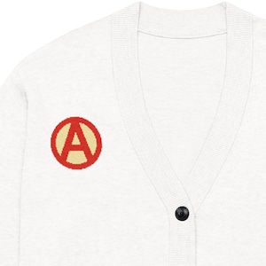 Può includere: Un cardigan grigio chiaro con scollo a V e bottoni neri. Il maglione presenta una toppa rossa e gialla sul petto sinistro. La toppa ha un cerchio rosso con la lettera "A" gialla all'interno.
