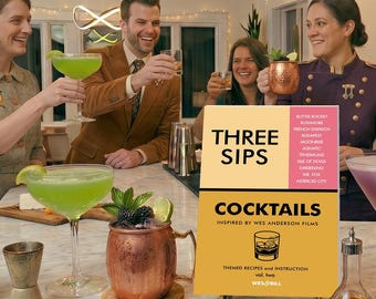 May include: A group of people toasting with cocktails at a home bar. The bar features a cocktail shaker, a copper mug, and a book titled "Three Sips Cocktails." Drinks include green and purple cocktails. The scene is well-lit with warm tones.
