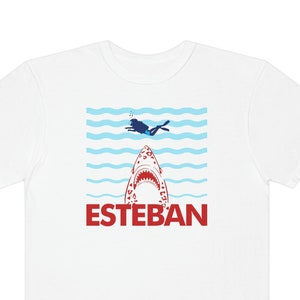May include: White t-shirt with a red shark graphic and the word "ESTEBAN" in red letters. A blue silhouette of a person diving is above the shark.