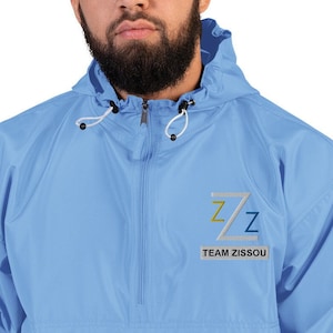 May include: Light blue Champion windbreaker jacket with a hood and a half-zip closure. The jacket features the text "Z/Z TEAM ZISSOU" in a rectangular patch on the chest. The jacket has a drawstring at the hood.