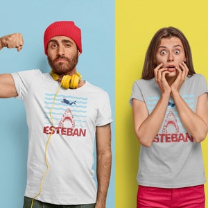 May include: Two people wearing gray and white t-shirts with a shark graphic and the text "Esteban". The person on the left is wearing a red beanie and has a yellow headphone around their neck. The person on the right is wearing a gray t-shirt and has their hands covering their mouth.