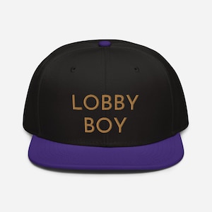 Peut inclure: Casquette de baseball noire et violette avec le texte "LOBBY BOY" brodé en or sur le devant.