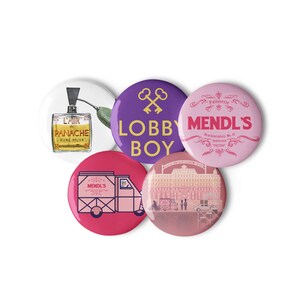 May include: Five colorful round buttons with different designs. One button features a purple background with a gold cross and the text "LOBBY BOY". Another button features a pink background with the text "MENDL'S Patisserie Brucknerplatz, No. 12 NEBELSAR". A third button features a white background with a drawing of a bottle of perfume labeled "LAIR de PANACHE PURE MUSK". A fourth button features a pink background with a drawing of a delivery truck labeled "MENDL'S". The last button features a pink background with a drawing of a building.