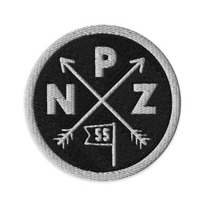 Könnte beinhalten: Schwarzer und weißer gestickter Patch mit einem kreisförmigen Design. Die Mitte des Kreises zeigt zwei gekreuzte Pfeile mit einer Flagge unten. Die Buchstaben "NZ" befinden sich links und rechts der Pfeile, und der Buchstabe "P" befindet sich über den Pfeilen.